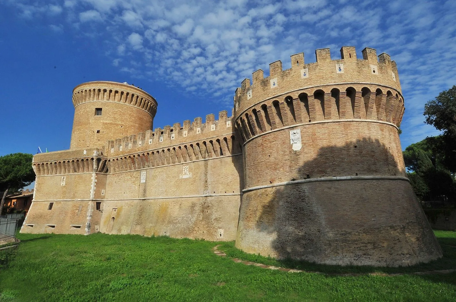Il Castello di Giulio II a Ostia Antica sotto un cielo azzurro a nuvole sparse: le mura in mattoni coronate da merli e beccatelli, il torrione circolare in primo piano e il mastio cilindrico sullo sfondo, con il prato davanti