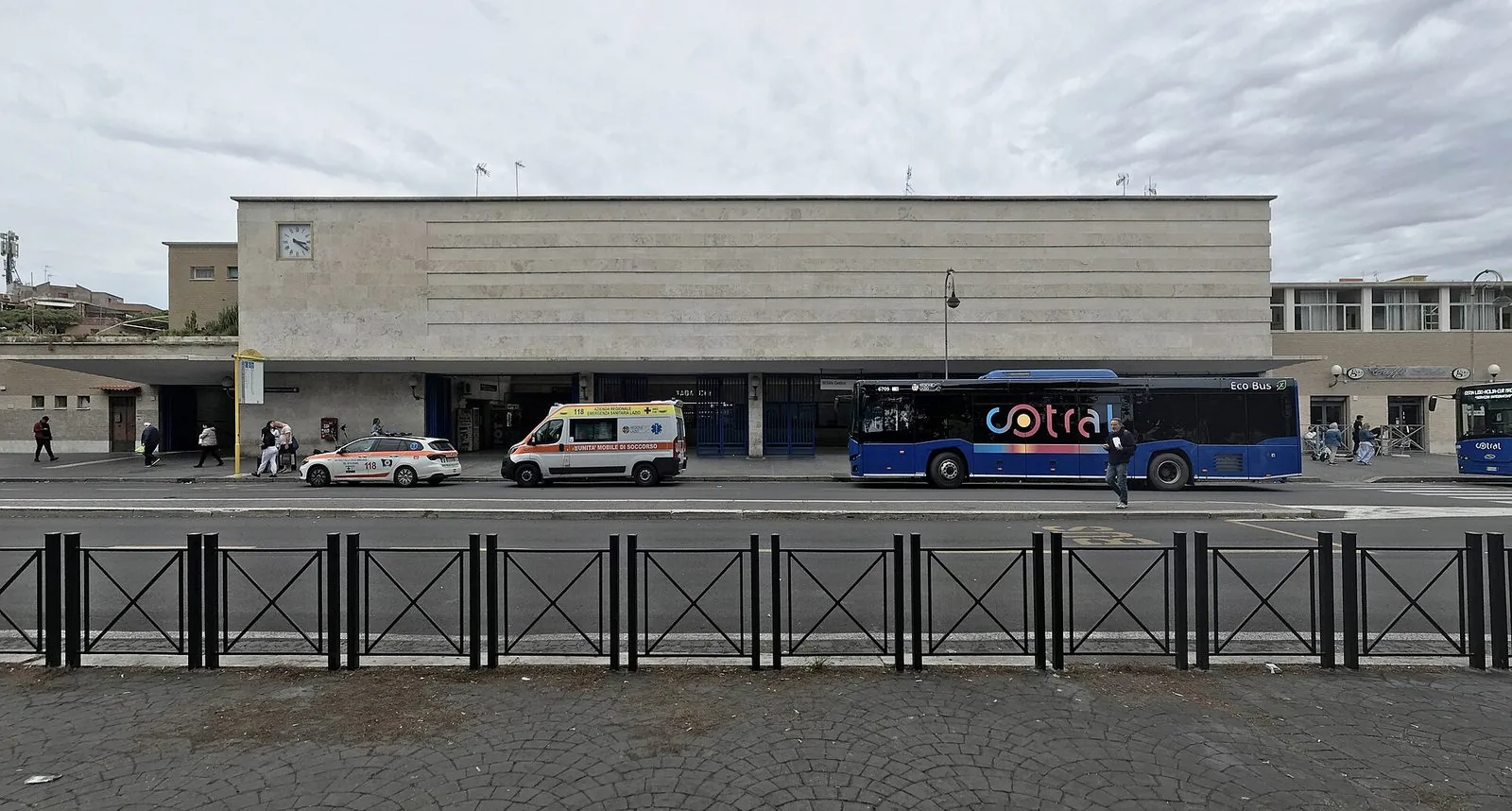 La facciata della stazione Lido Centro della ferrovia Roma–Lido, un lungo edificio rivestito in travertino con l'orologio in alto a sinistra, un autobus blu e un'ambulanza ferma davanti all'ingresso