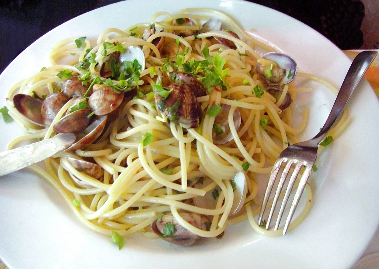 Un piatto bianco di spaghetti alle vongole con prezzemolo fresco tritato, un coltello e una forchetta appoggiati sul bordo