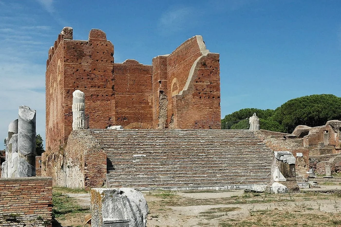 Il Capitolium di Ostia Antica: l'alto tempio in mattoni rossi con la grande scalinata di accesso, colonne spezzate di marmo in primo piano e pini sullo sfondo sotto un cielo azzurro