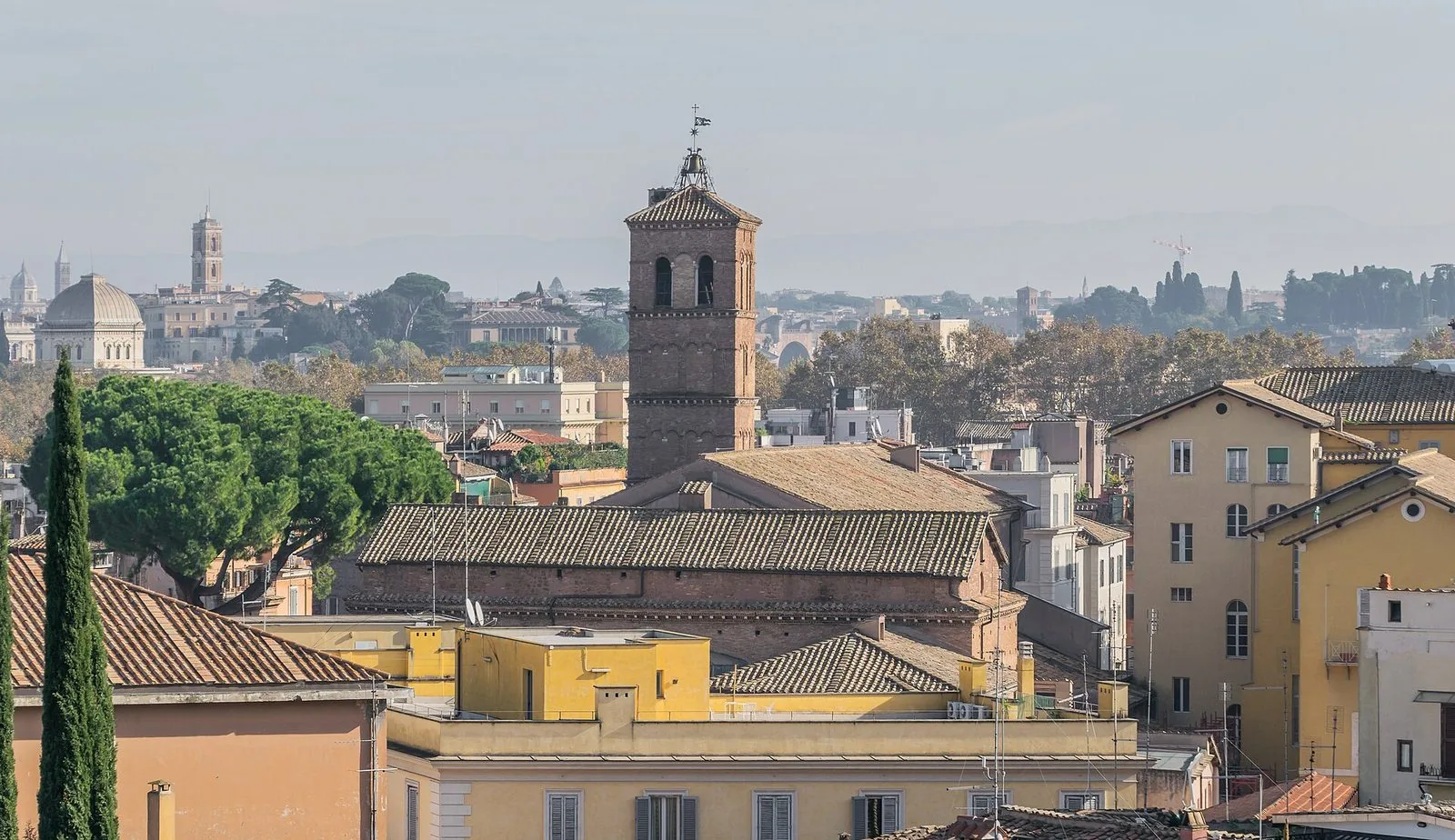 I tetti di Trastevere visti dall'alto con il campanile romanico in mattoni di Santa Maria in Trastevere al centro, pini e cipressi a sinistra e le cupole di Roma all'orizzonte nella foschia