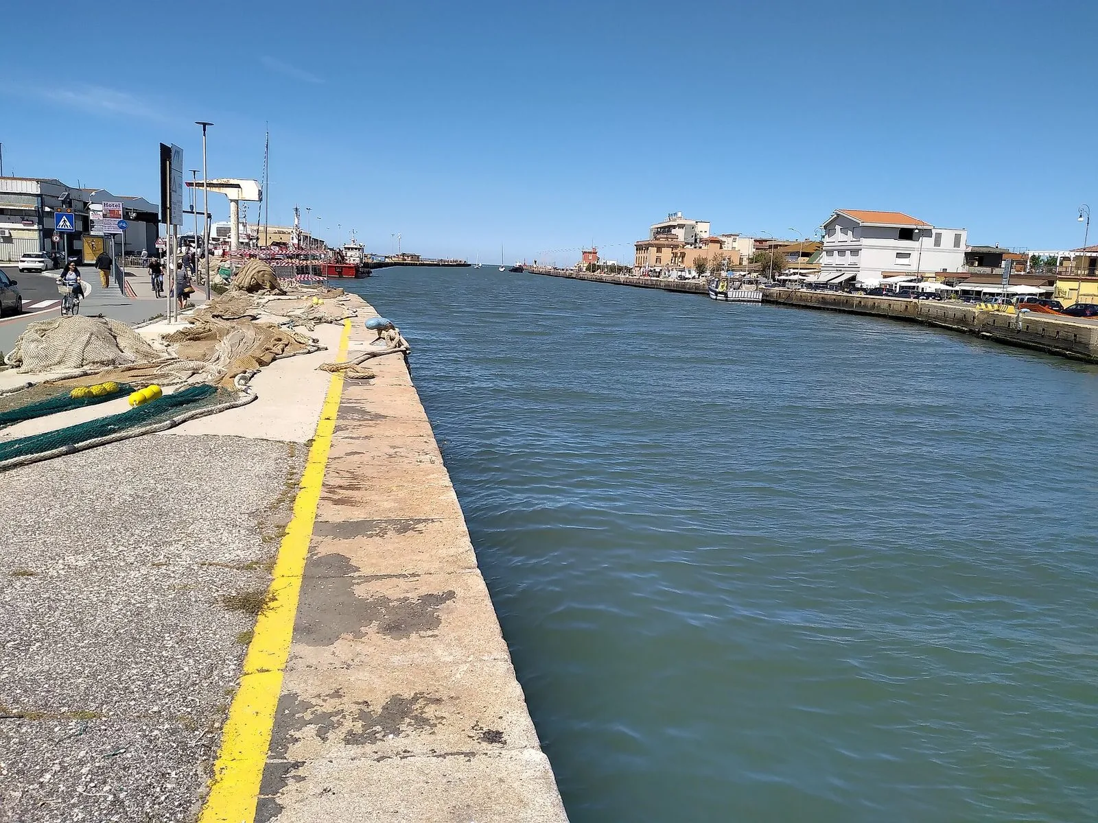 Il porto canale di Fiumicino in una giornata limpida: la banchina con la riga gialla e le reti da pesca stese sulla sinistra, l'acqua verde del canale al centro e sulla riva opposta case basse e locali affacciati sull'acqua