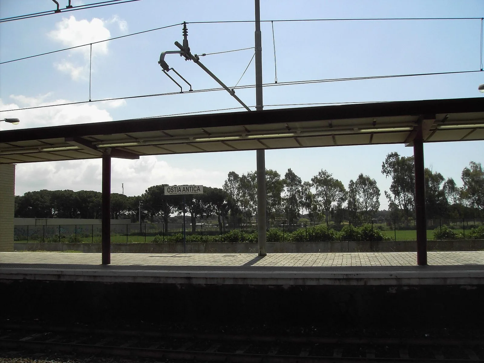 La banchina della stazione Ostia Antica della ferrovia Roma–Lido, con la pensilina, il cartello con il nome della fermata e sullo sfondo pini domestici ed eucalipti sotto un cielo azzurro