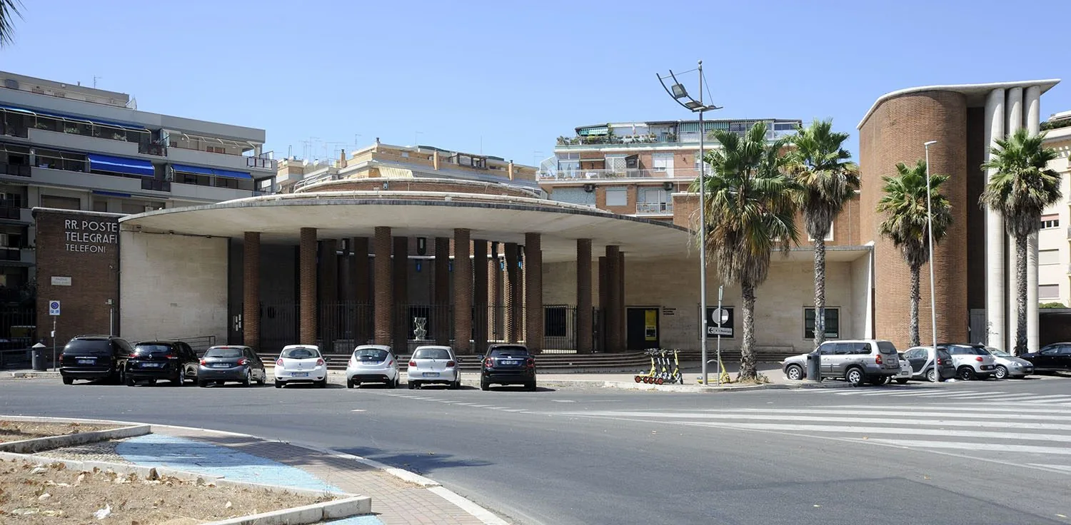 Il Palazzo delle Poste di Ostia: il grande portico curvo sorretto da colonne in mattoni, la scritta RR. Poste Telegrafi Telefoni a sinistra e la torre cilindrica in mattoni con colonne bianche a destra, con palme e auto parcheggiate davanti