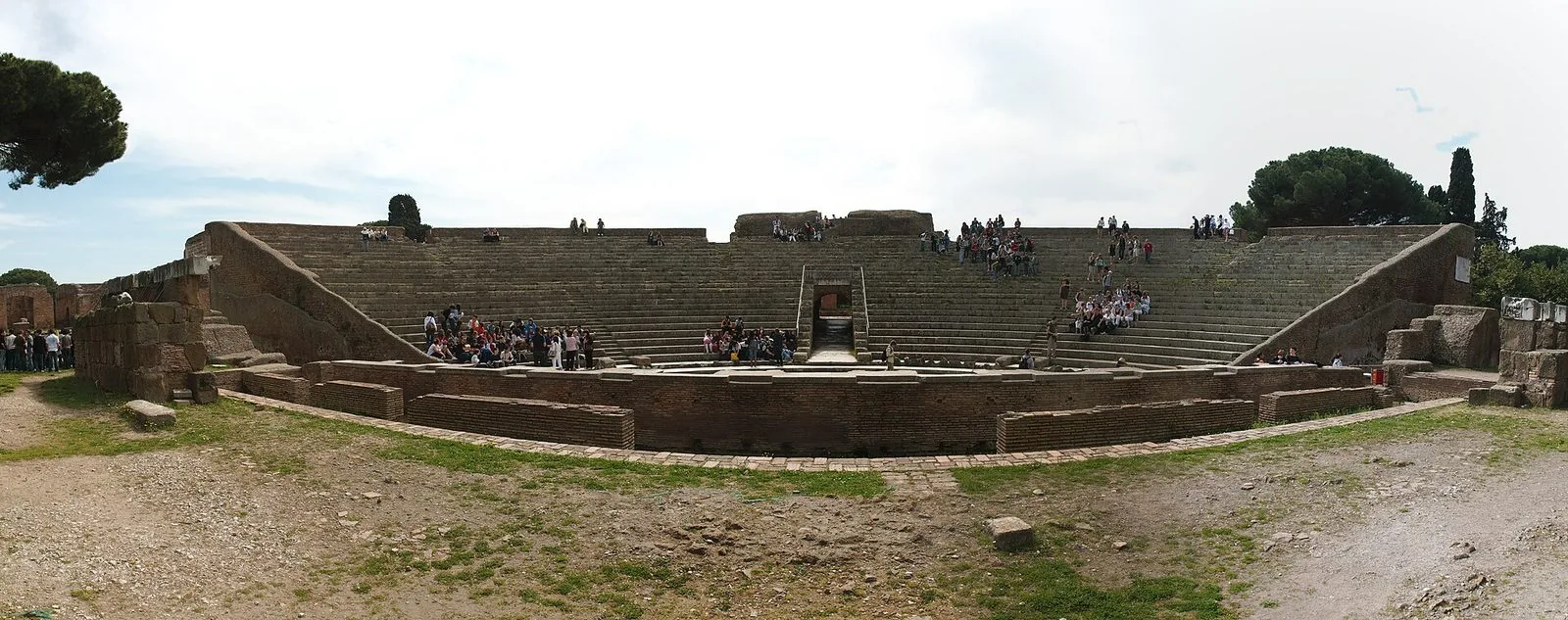 Veduta panoramica del Teatro romano di Ostia Antica: la cavea semicircolare a gradinate in pietra, il muro dell'orchestra in mattoni e piccoli gruppi di visitatori seduti sui gradini, con pini sullo sfondo