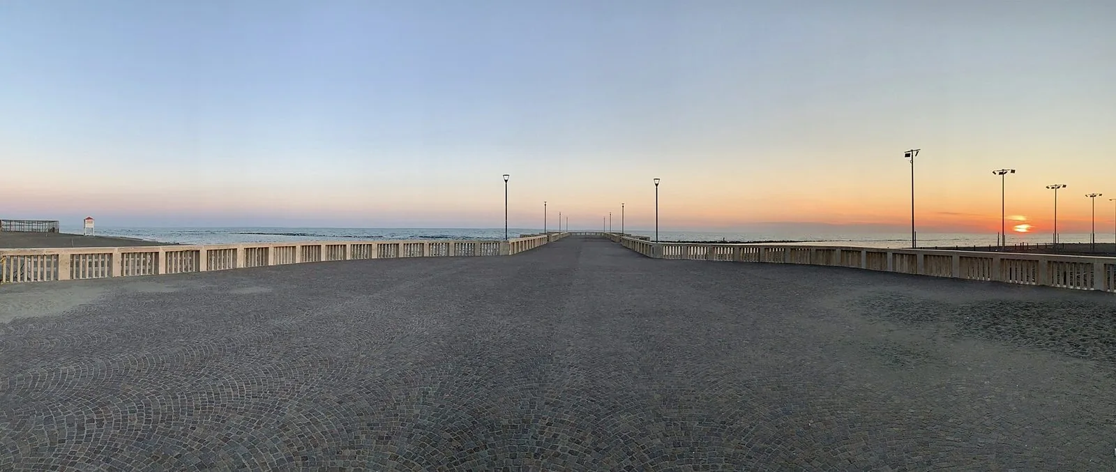 Il Pontile di Ostia al tramonto in una foto panoramica: la piazza lastricata a sampietrini, le balaustre chiare e i lampioni che accompagnano lo sguardo verso il mare, con il sole che scende all'orizzonte a destra
